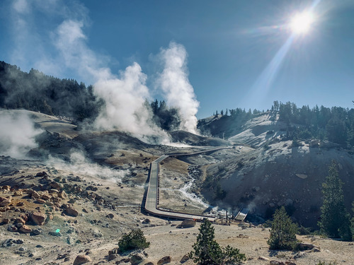 White clouds of steam rise from a hydrothermal area. A boardwalk passes through the center toward a setting sun in the top right. 