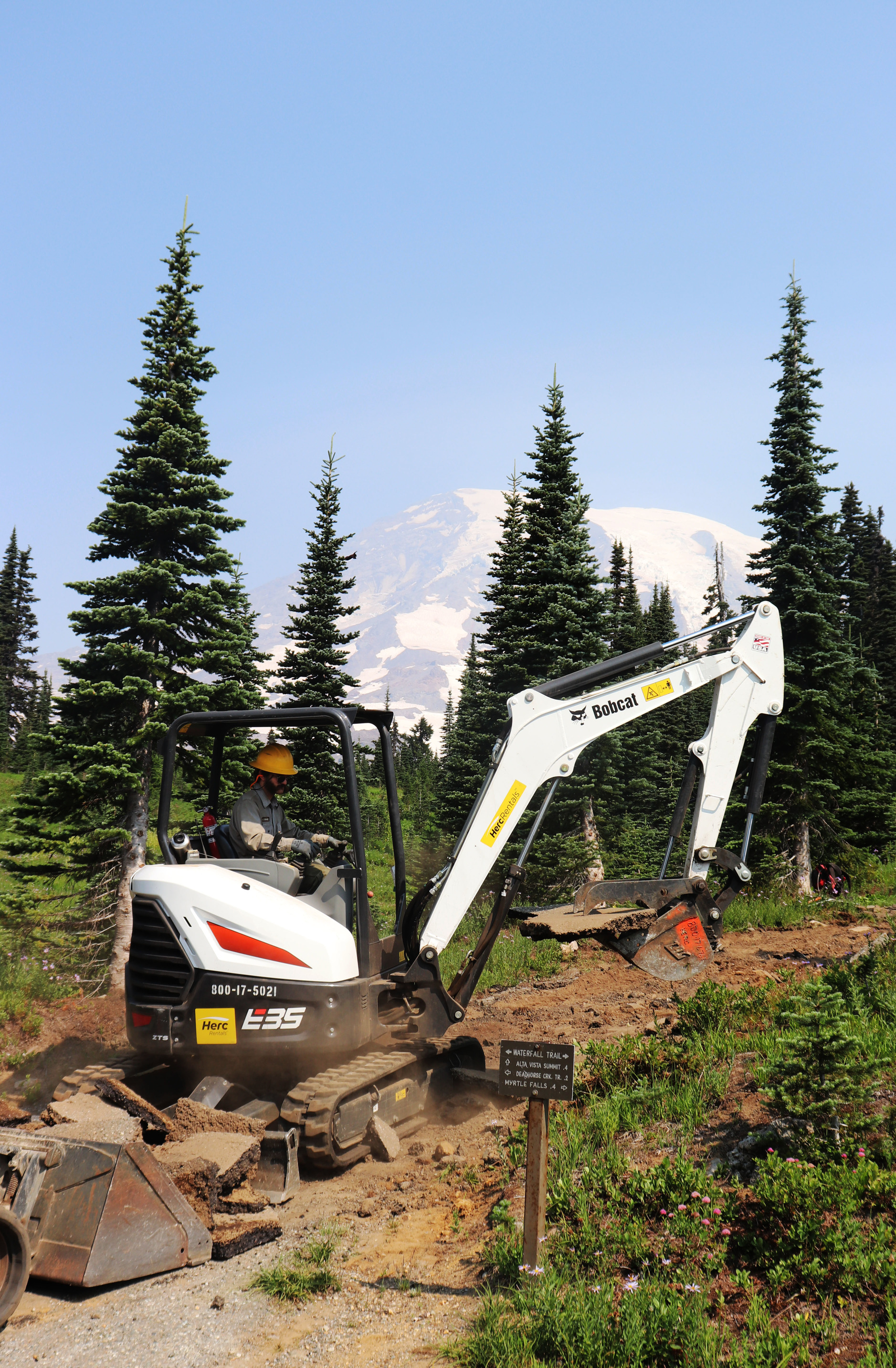An employee operates an excavator on a dirt trail with Mount Rainier looming in the background. 