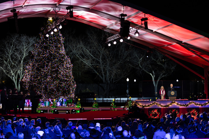 The president and first lady stand at a podium as they light a large 40 foot tall Christmas tree behind a stage. 