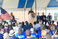 Uniformed NPS employee speaks from behind a podium to a large crowd seated under a big blue and white tent. Arrowhead logo on the podium reads, 