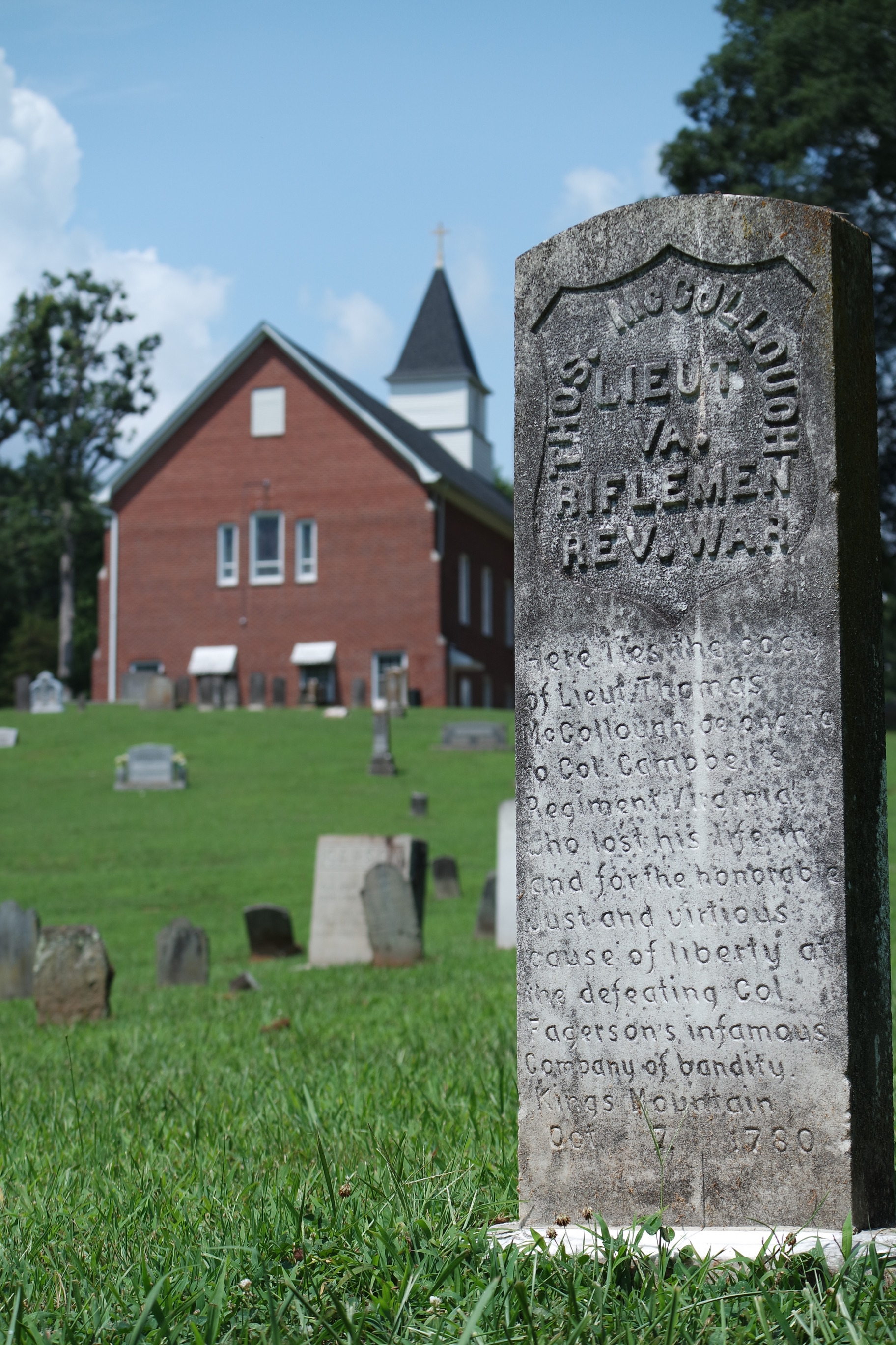 A tall grey headstone in a green grassy cemetery in front of a red brick church. The top of the stone reads "Thos. McCullough. Lieut. VA. Riflemen. Rev. War." The lower part of the stone reads "Here lies the body of Lieut. Thomas McCollough, belonging to Col. Campbell's Regiment Virginians who lost his life in and for the honorable just and virtuous cause of liberty at the defeating Col. Fagerson's infamous Company of bandity. Kings Mountain, Oct. 7. 1780."