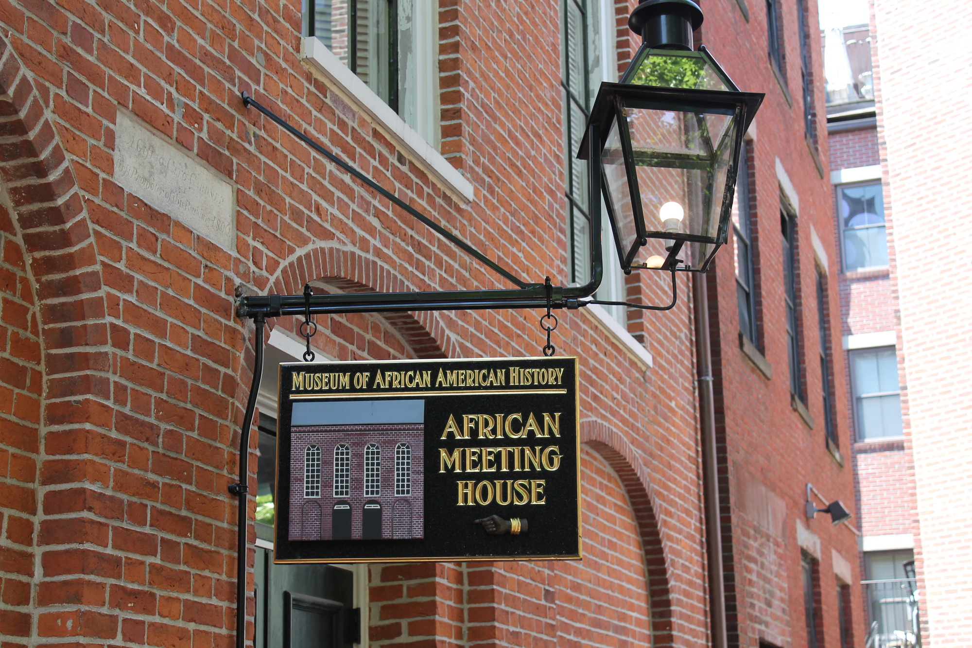 An outdoor image take from the street. The African Meeting House is made of dark red bricks and is only partially shown. A sign post is in the center of the image as it extends out parallel to the street below. The sign hanging from it is rectangular, made of wood, and painted black. It shows an image of the building with the words Museum of African American History above and African Meeting House beside it. There is also a hand pointing to the left underneath those words. The hand is of an African American. An old fashion light is at the very end of the sign pole.