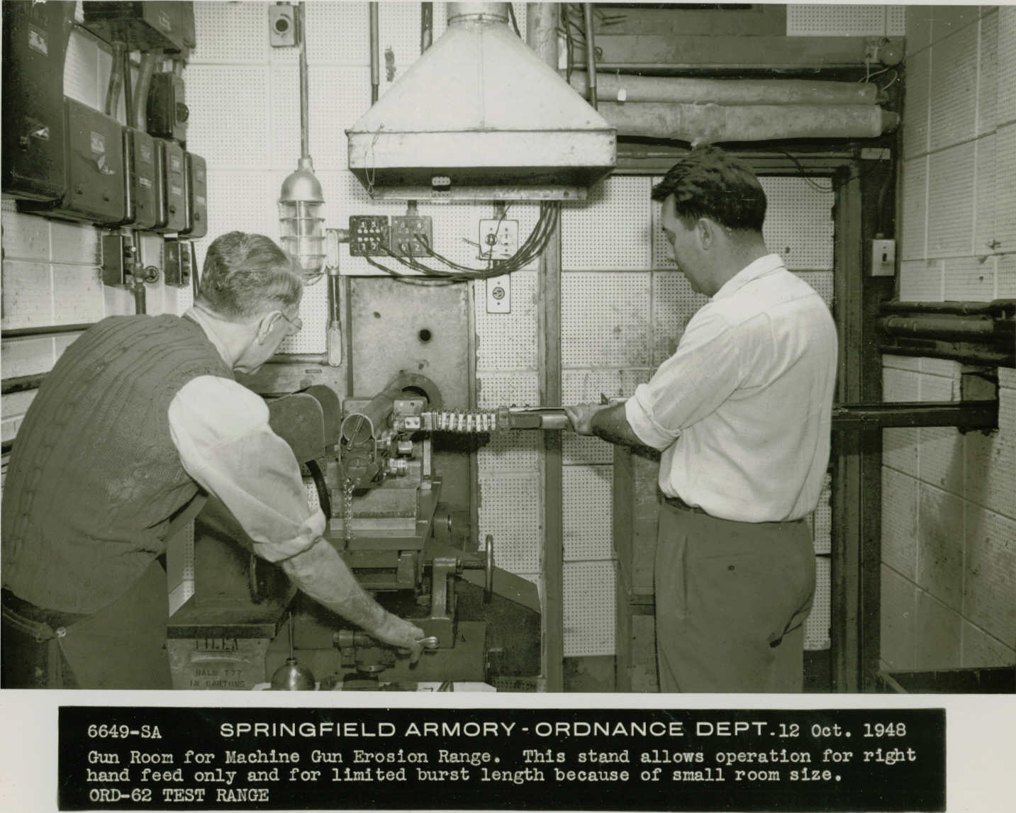 Black and white photo of two men working on a piece of machinery in a small room. One man wears an apron. Text under the photo reads, “6649-SA Springfield Armory – Ordnance Dept. 12 Oct. 1948, Gun Room for Machine Gun Erosion Range. This stand allows operation for right hand feed only and for limited burst length because of small room size., ORD-62 TEST RANGE.”