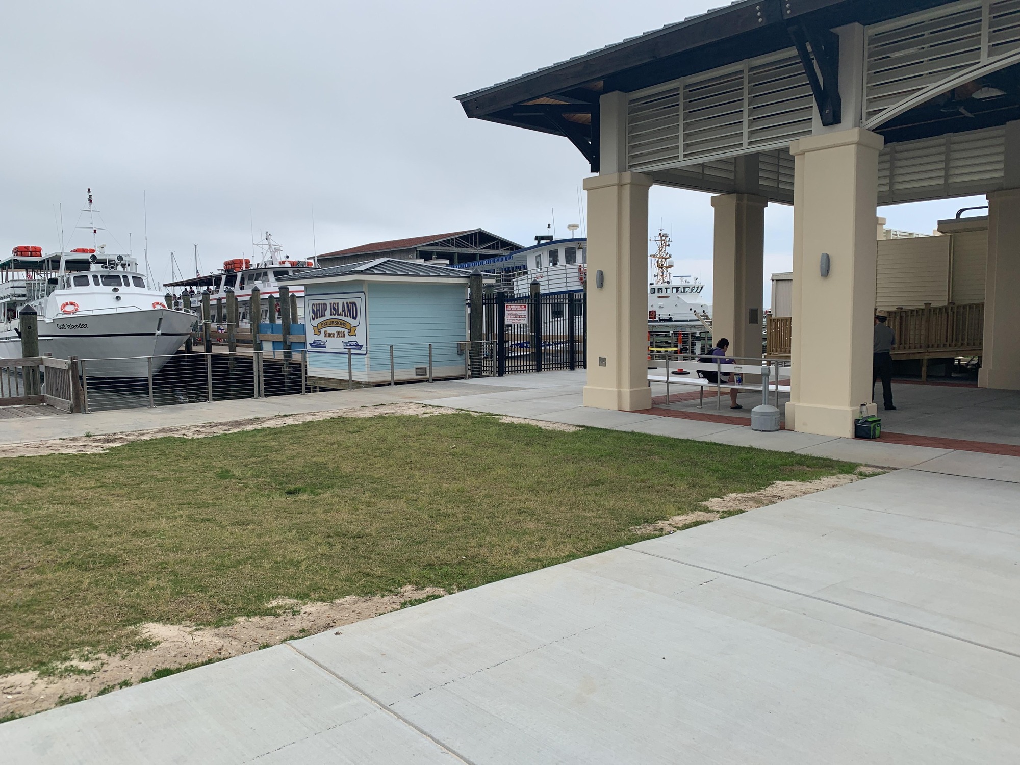 Cement building with metal benches beneath. Boats docked near a pier in the background.