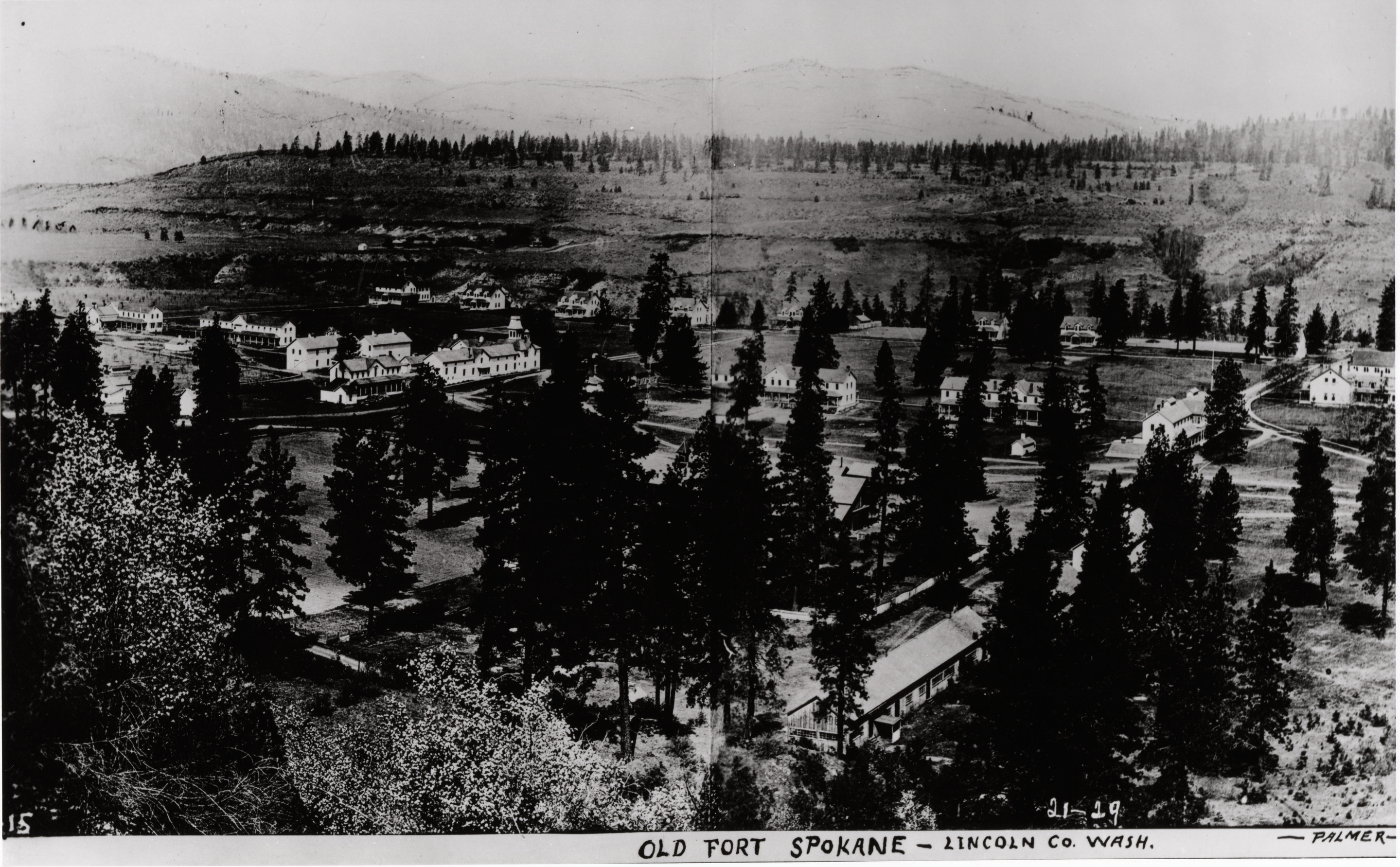 Black and white photograph of many buildings in a wooded valley