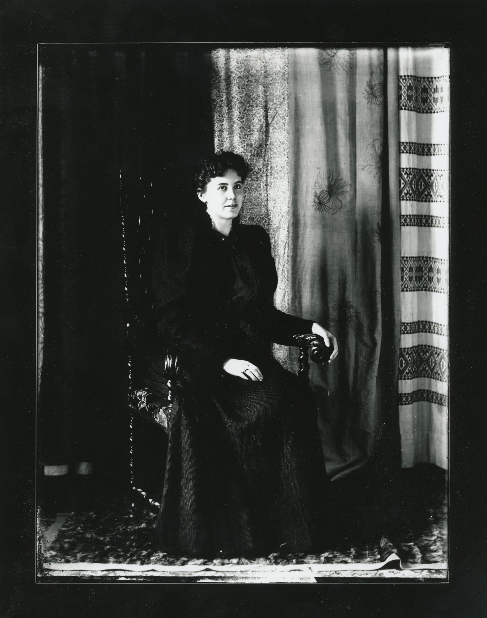 A white woman in dark clothing with dark hair sits in a chair on a carpet in front of a background drape.