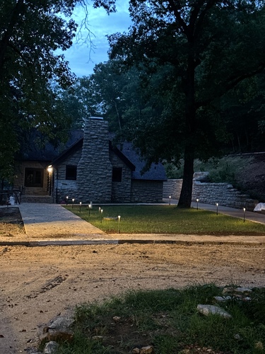 An evening photograph of a stone building with a large stone chimney facing the viewer, framed by trees on either side. Concrete walkways leading to the building are highlighted by path lights.