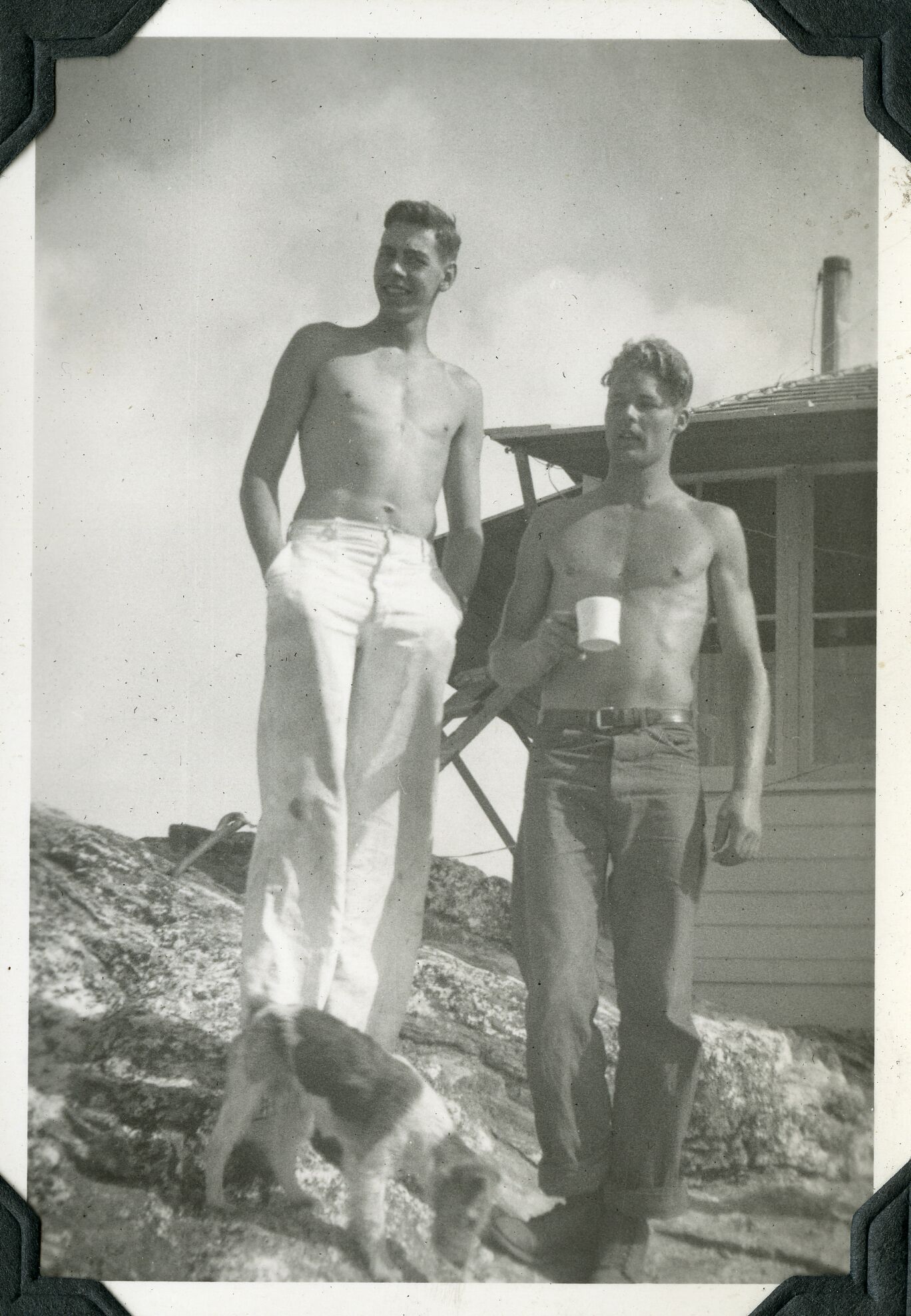 Two shirtless young white men standing with a terrier on rocky terrain in front of a wooden structure.