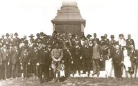A black and white photo of a crowd of people looking at the camera in front of the base of a statue. 
