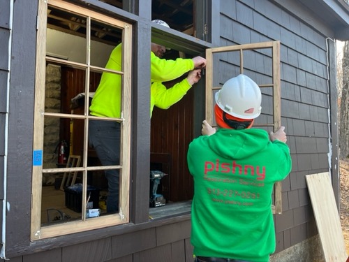 Two workers are setting an unstained window pane into a divided window frame in a dark brown stained shingle-sided building. One worker, facing away, wearing a white hard hat ack a green hoodie that reads "Pishny Restoration Services" in red, stands outside the building, holding a pane in place while another worker, visible through the window frame and another pane already in place, wearing a white hard hat and fluorescent yellow sweatshirt, attaches a hinge to the pane held by the first worker.