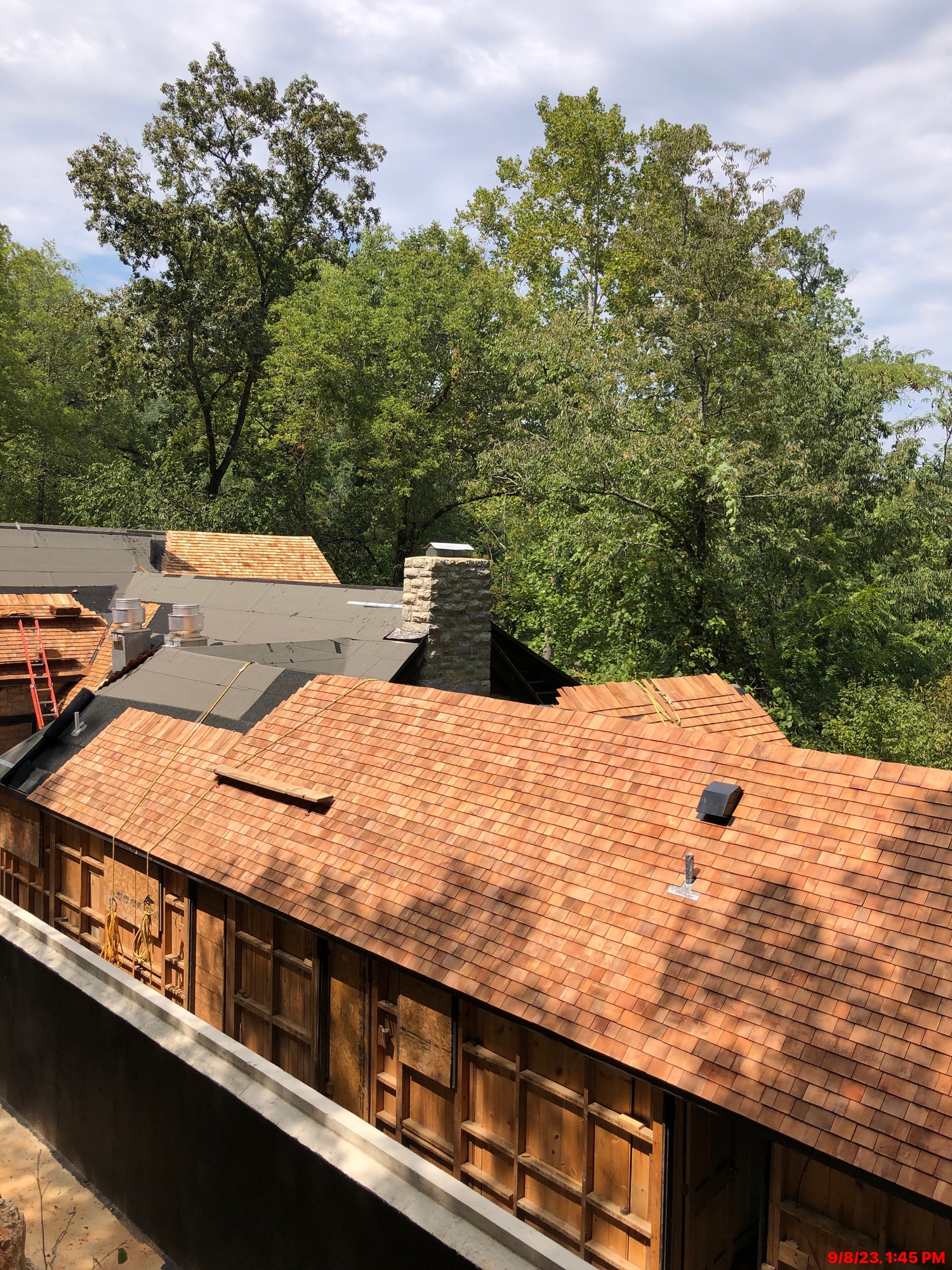 The roof of a long building that changes direction at right angles several times as it progresses into the distance is in the process of being covered with new cedar shingles. A group of green trees is visible above the roof in the distance with a cloudy sky above.