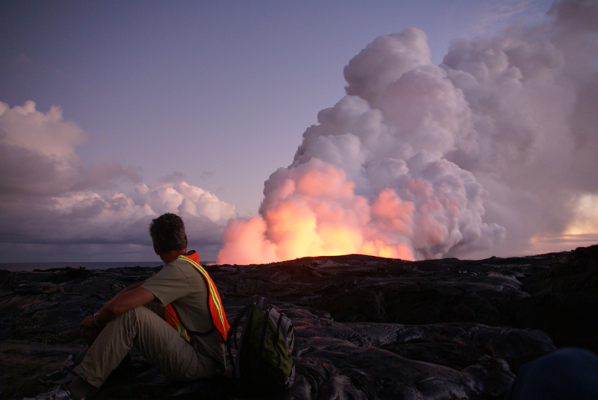 A person sits looking at a glowing plume of smoke in the distance