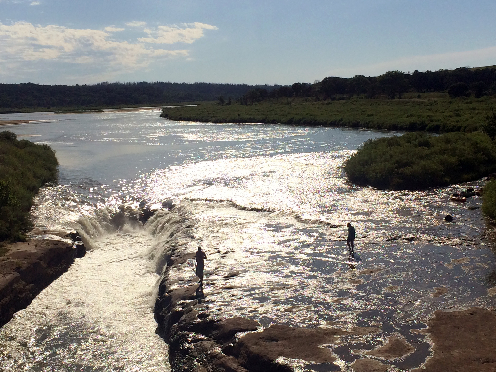 Two people walk along a sunlit wet rocky bank next to a dramatic rapid in a river.