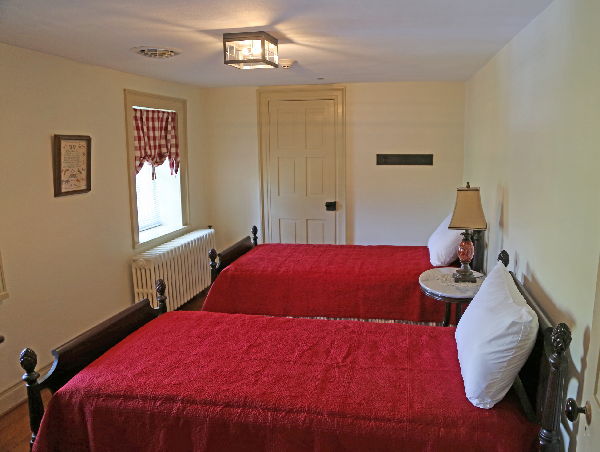 Two twin beds have matching red quilts and a single white pillow. In between the beds is a circular marble topped table with a lamp on top. The window features a red and white checkered pattern. A piece of art hangs in between the windows on the wall. 