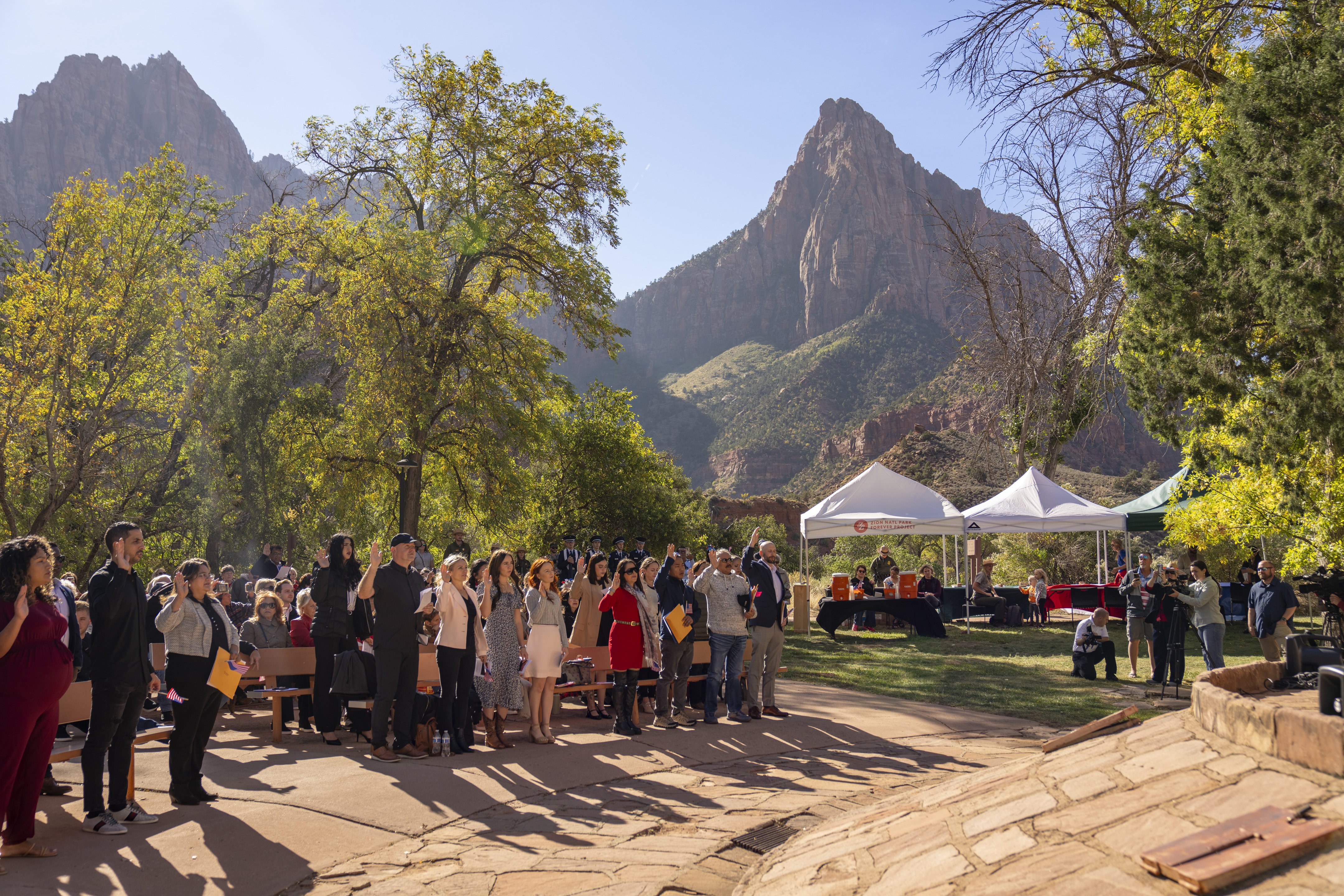 Rows of people stand with a raised arm to recite the Oath of Affirmation. In the background are two white shade structures and a distant sandstone peak. 