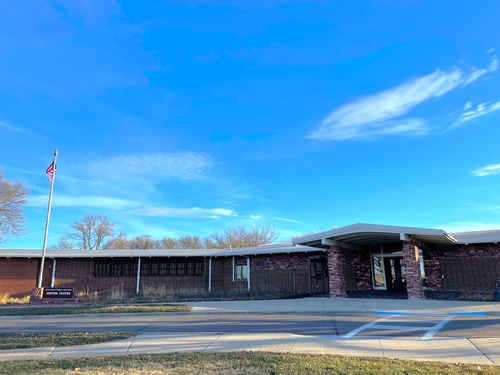 A long, low 1-story building under blue sky, a sidewalk and grass in the forefront and an American flag to the left. 