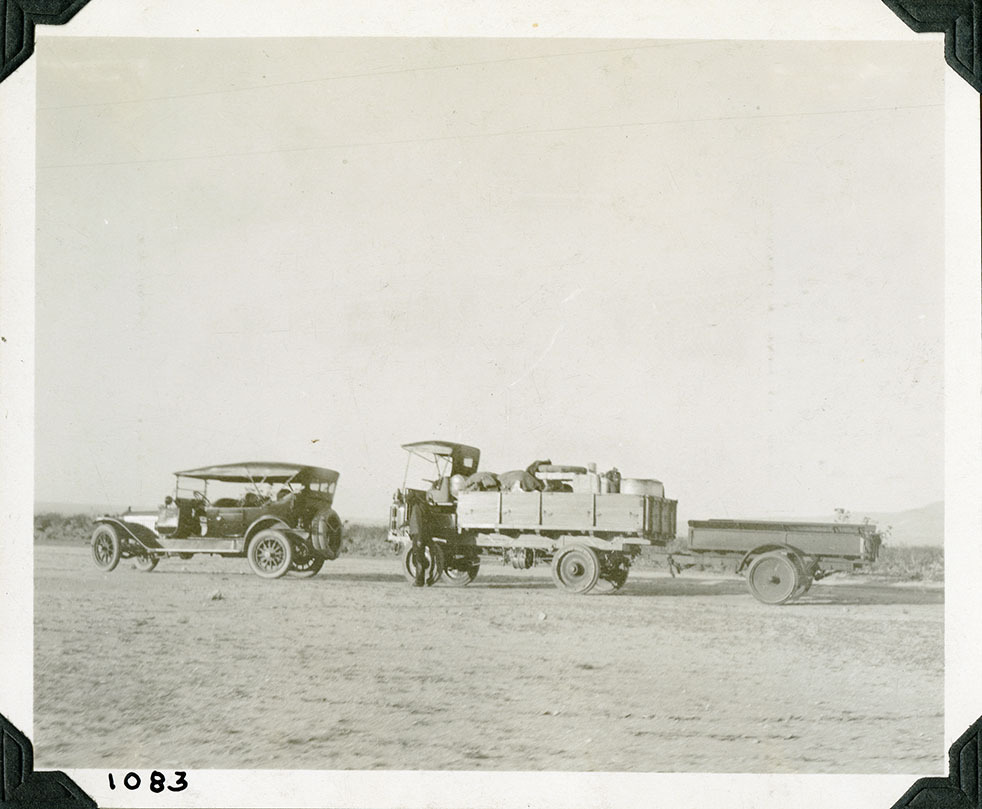 This is an historic black and white photograph from the Scotty's Castle Historic Photograph Collection, Death Valley National Park of a passenger car and a heavy-duty truck hauling supply trailer. Car is two door and open with canopy. Truck bed fully of various supply, some covered with canvas. Long and low-profile load in trailer. Parked on flat dirt area with sparce vegetation in background. Number in black ink in lower left corner.