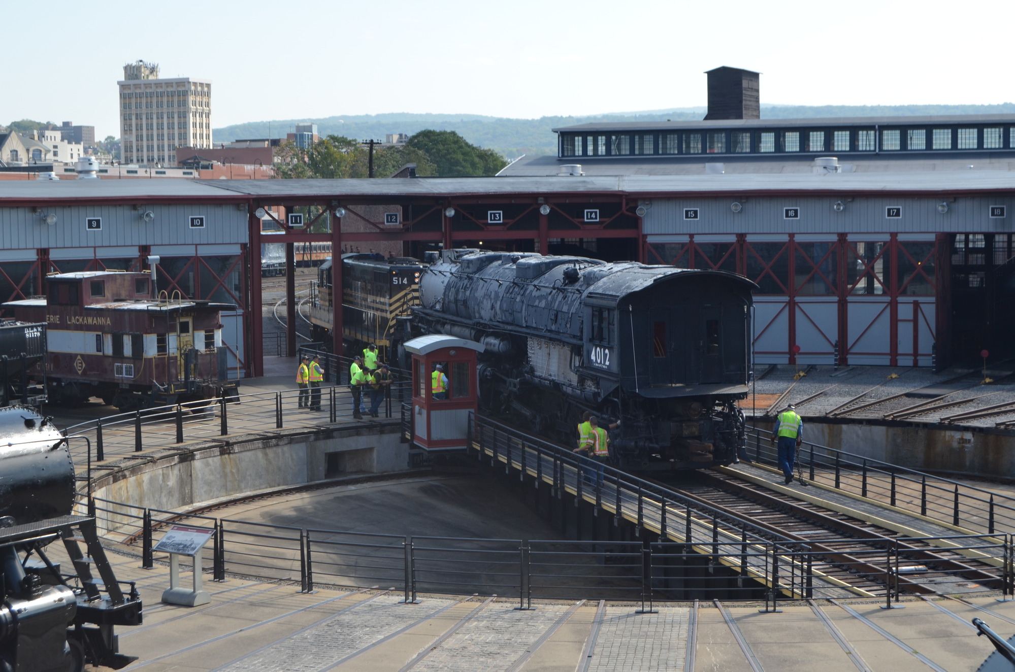 Elevated exterior view of a large train on a turntable with 8 people looking on. A second train is in a threshold moving towards the turntable.