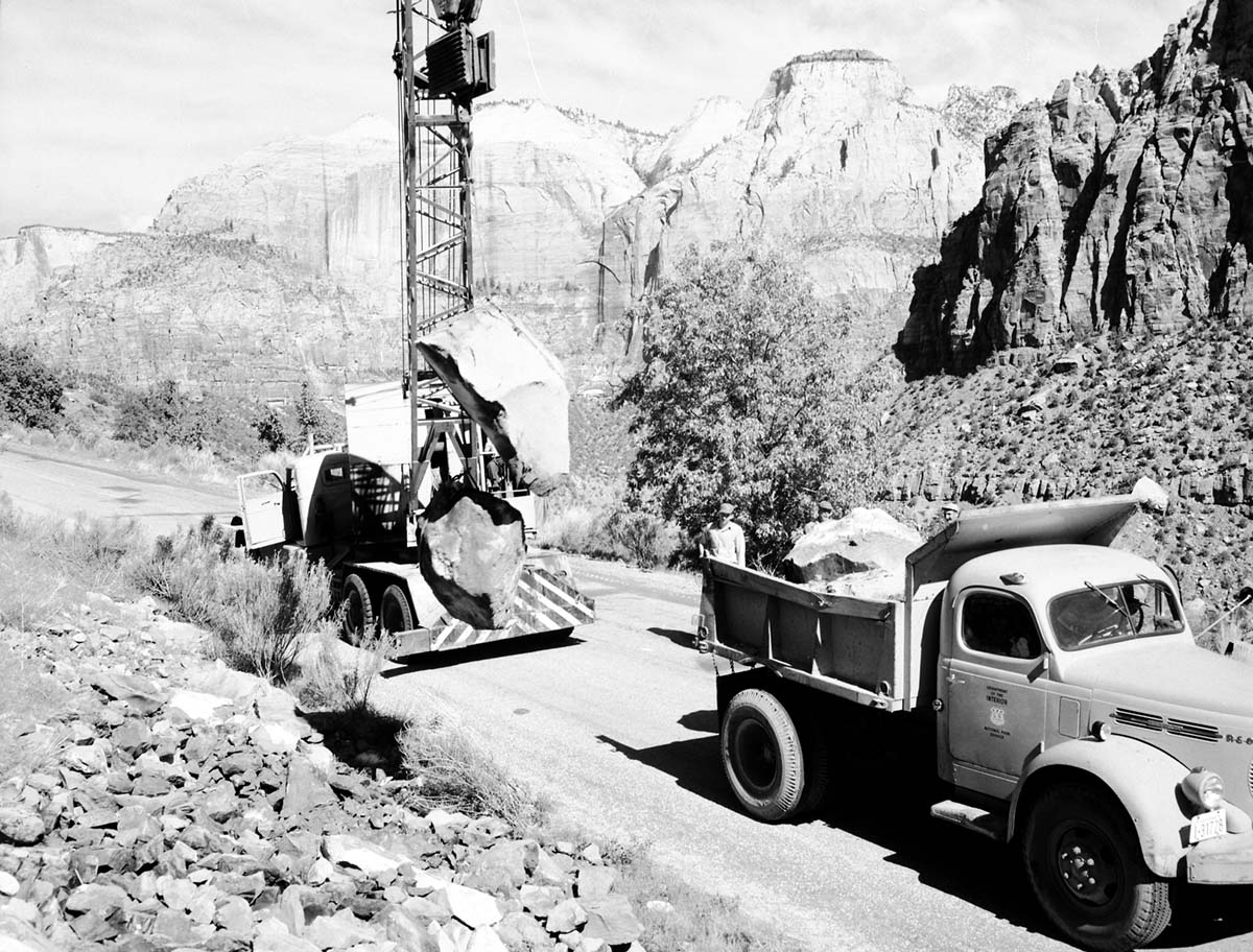 A dump truck and crane truck hauling rocks for revetment work to control Virgin River flood damage. Workers standing on the back of the dump truck. Mount Spry (right), Streaked Wall and Sentinel (center).