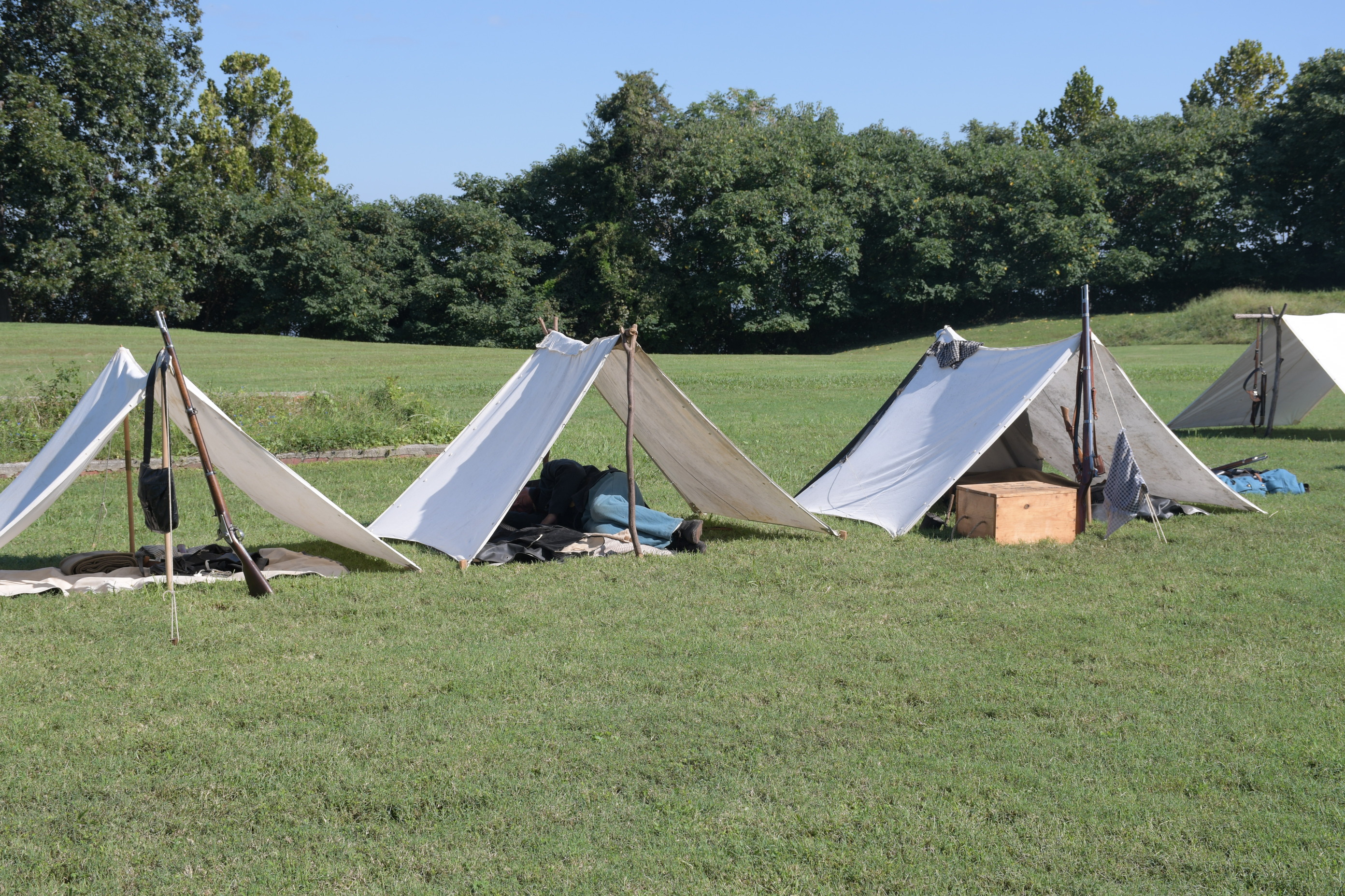 A man wearing a US army uniform sleep in the middle of three white a-frame tents.  Clothing and rifles lean against the tent poles and guidewires of two of the tents.