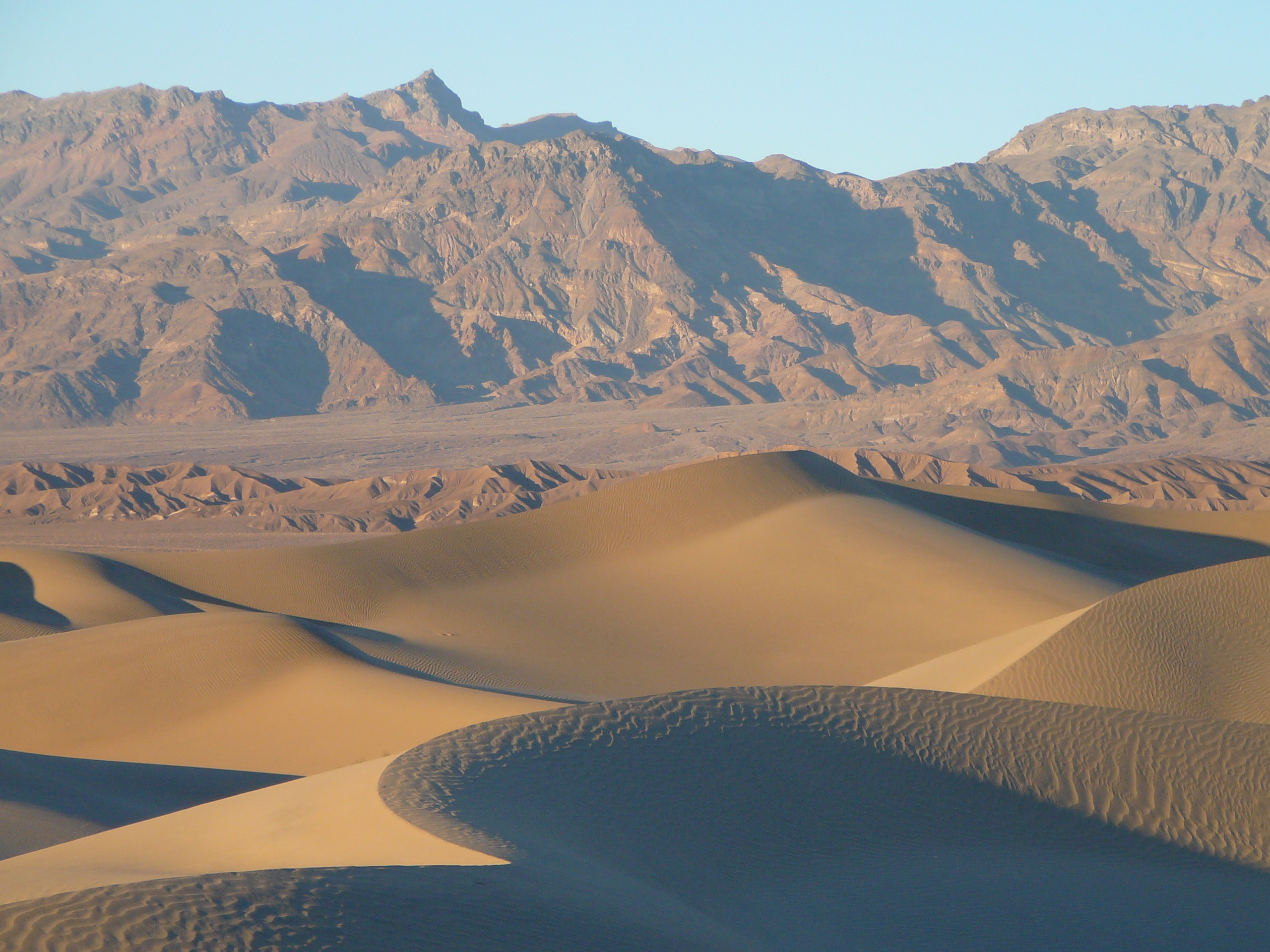 Tan sand dunes, with tall brown mountains in the background.