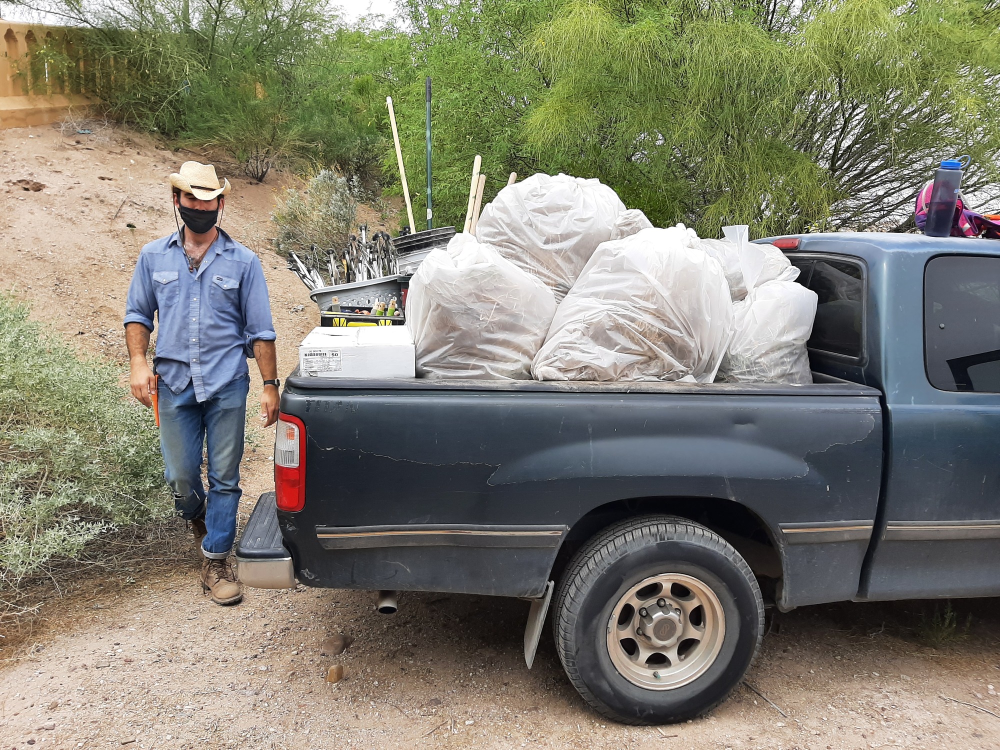 A blue truck is filled with full garbage bags and picker uppers with a person in boots and a cowboy hat walking behind