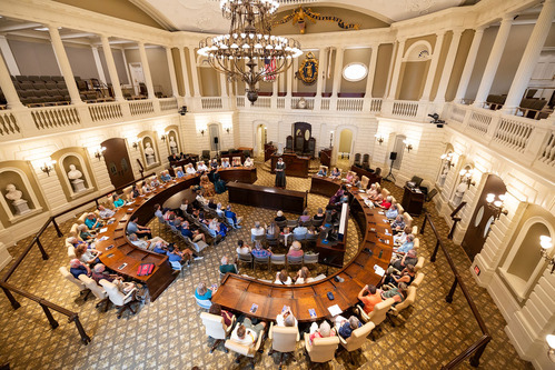 A circular chamber. Looking from the upper gallery onto the floor, which has two semi-circles of seats. Seating is in the middle of the two semi-circles. Actors are sitting towards the head of the room, with one actor standing behind a podium.