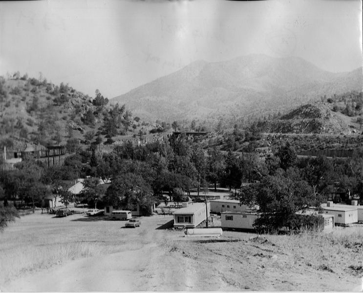 Forested hillsides with several white trailers in the foreground, on the edge of an open grassy area. 