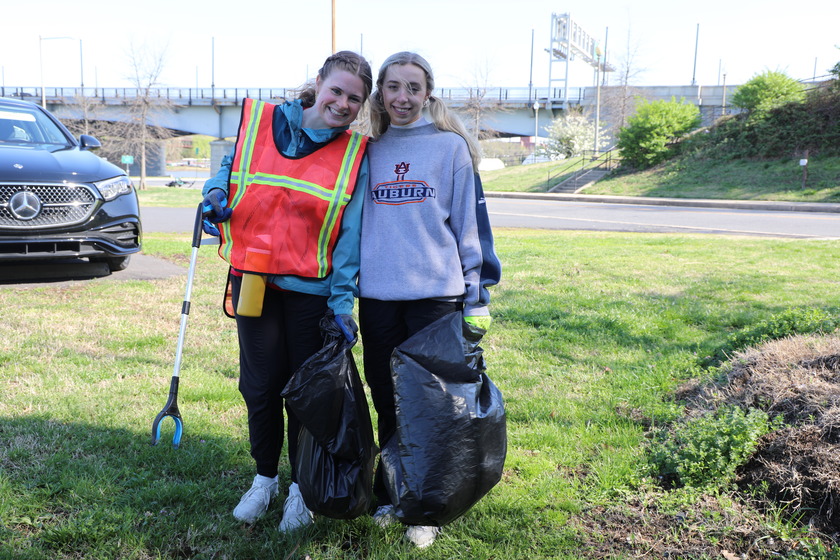 Two young women stand side-by-side in a grassy area, holding black trash bags full of collected litter. The woman on the left wears an orange reflective safety vest over a blue long-sleeved shirt, black pants, and white sneakers, holding a trash grabber tool. The woman on the right wears a grey long-sleeved shirt with "AUBURN" written on it, black pants, and white sneakers. Both are smiling at the camera. In the background, there's a black car, a road, and a bridge.