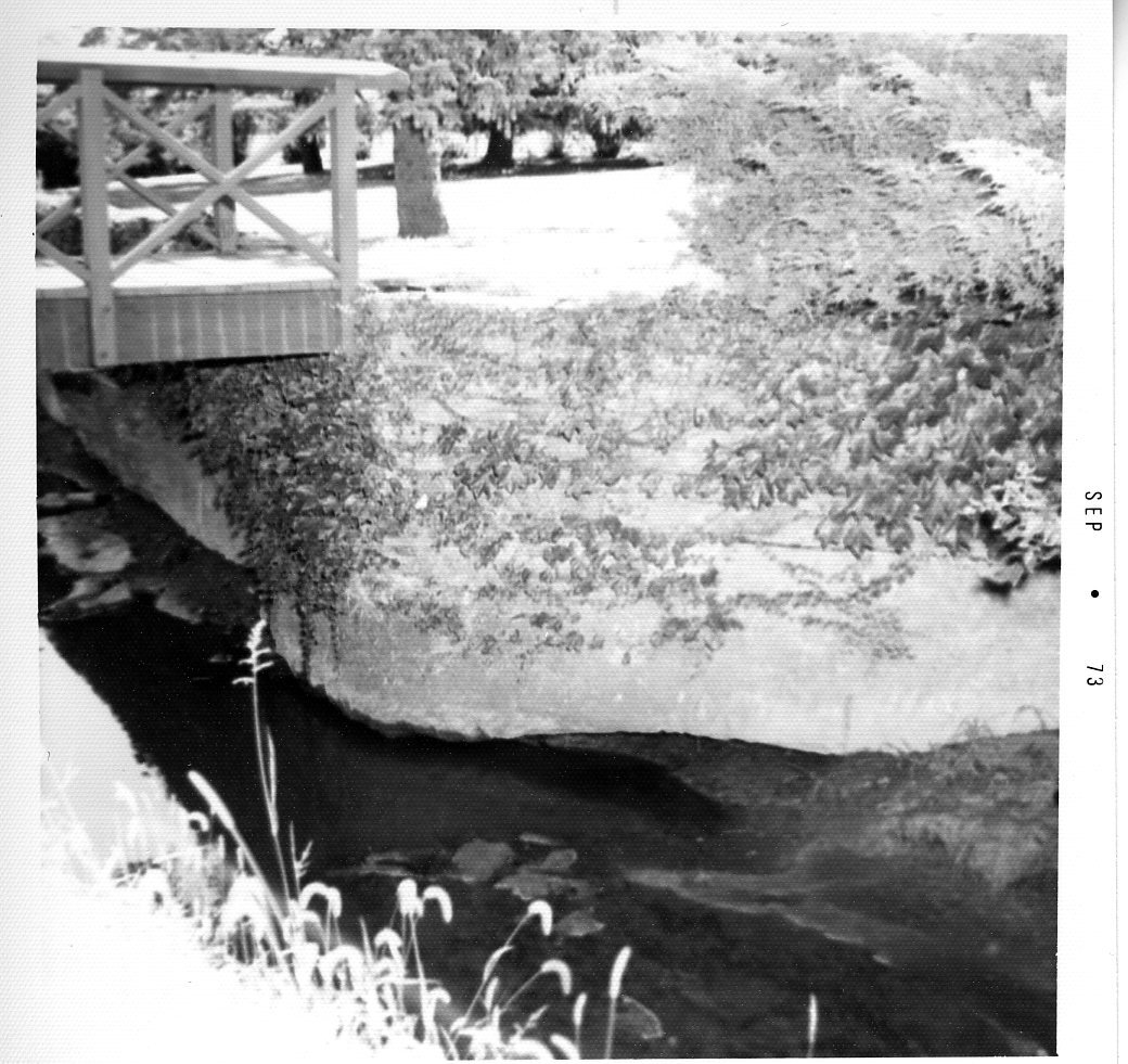 A 1973 photo shows erosion of a creek bank by the abutment of a truss footbridge.