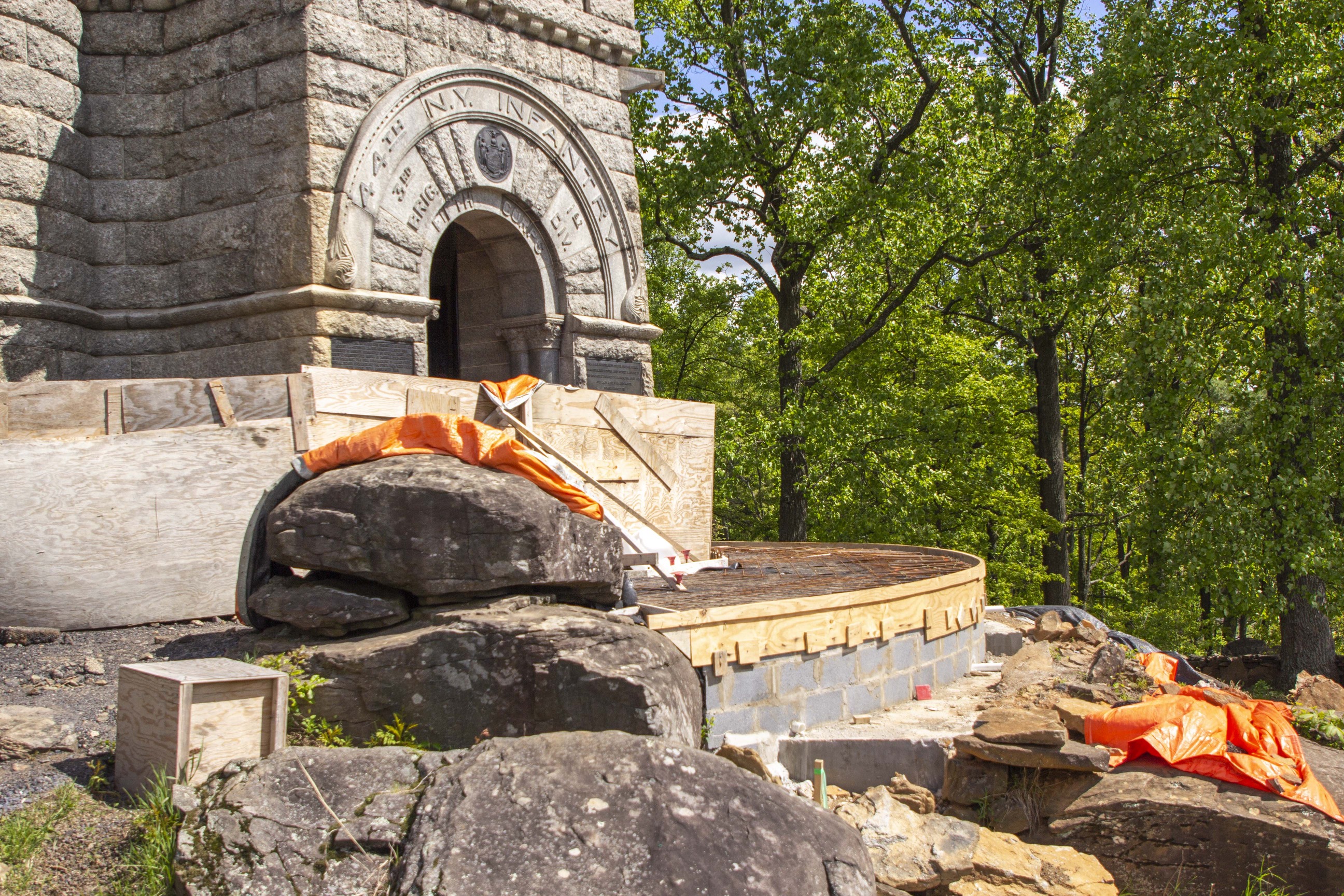 A stone wall with wood and rebar ready for concrete stands beside a large stone monument and large rocks with trees in the background.