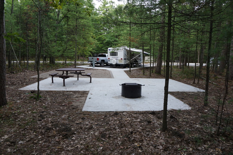 A view of a campsite in the forest of Sleeping Bear Dunes National Lakeshore with accessibility concrete pad, firepit and picnic table in foreground and camper with truck in background.