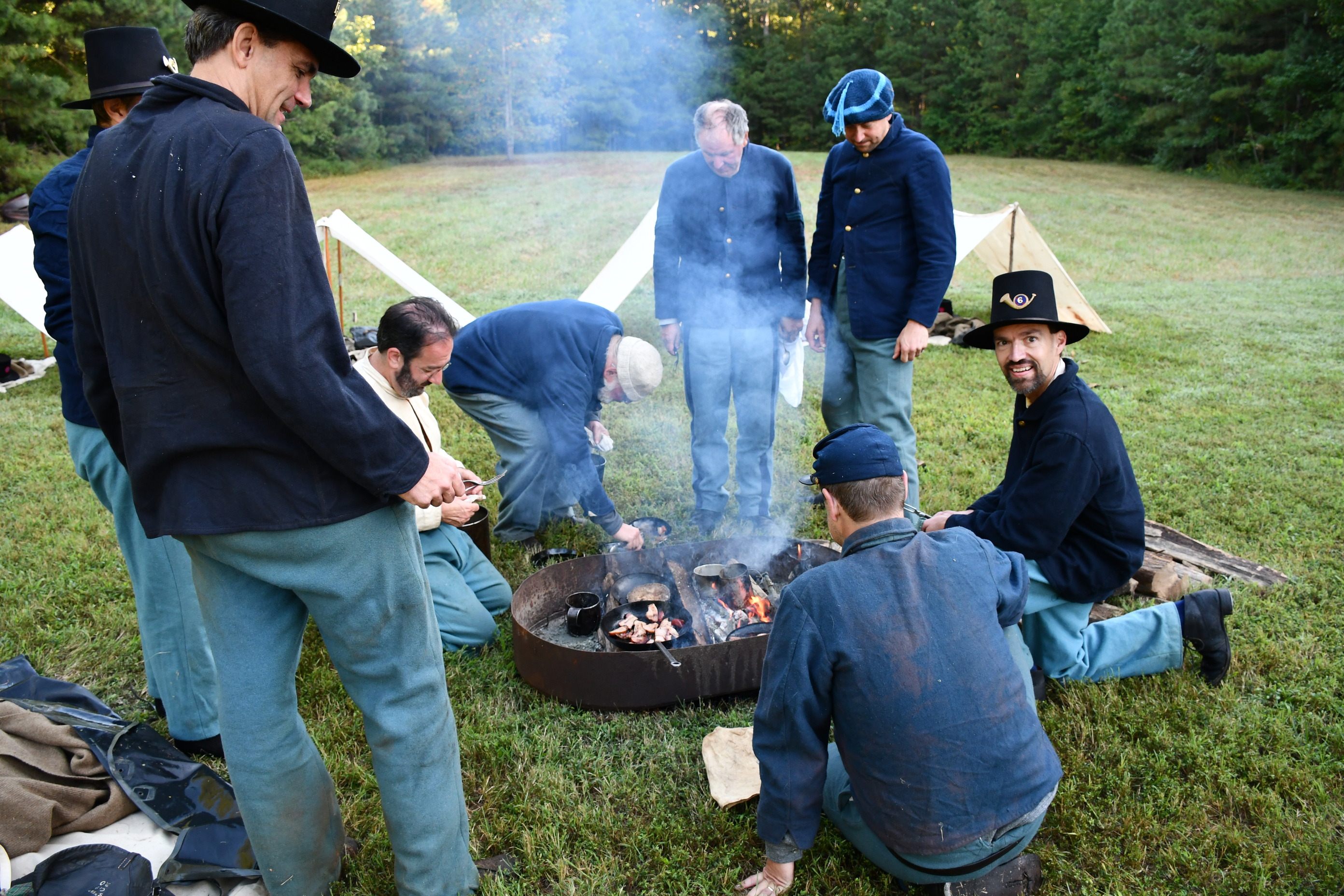 Eight white men in blue US Army unforms stand and kneel around an open fire pit cooking bacon, and boiling water in tin cups.