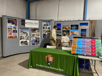 Inside a building two uniformed people in front of display wall with many photos, green arrowhead table in front.