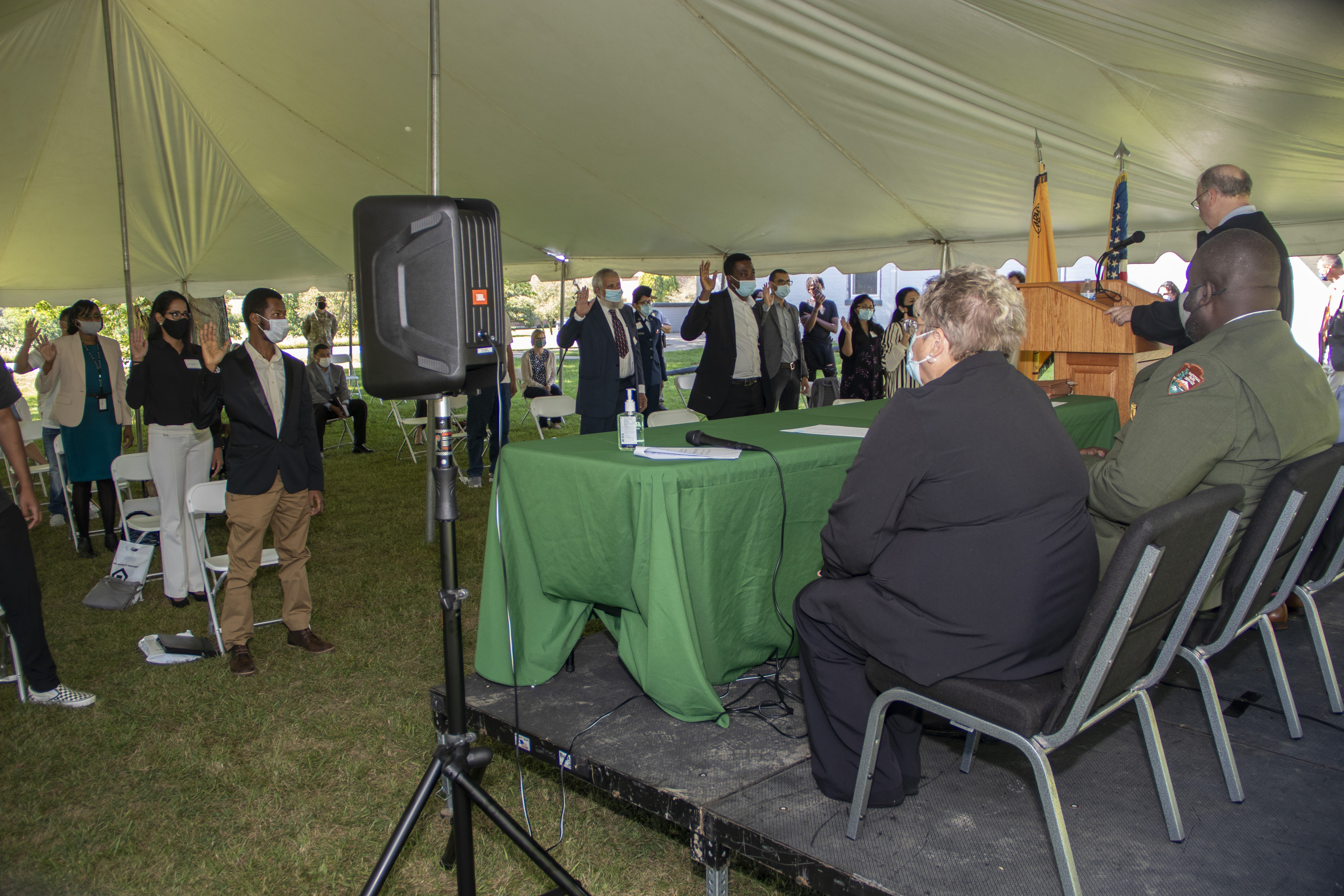 Several people standing and holding their right hands up in the air at shoulder height while two people sitting in chairs and one standing behind a podium face them from an elevated black stage behind a green-covered table.