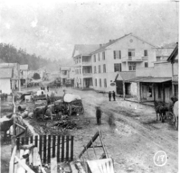 Mule drawn carts on an unpaved road in front of a large, three story hotel. 