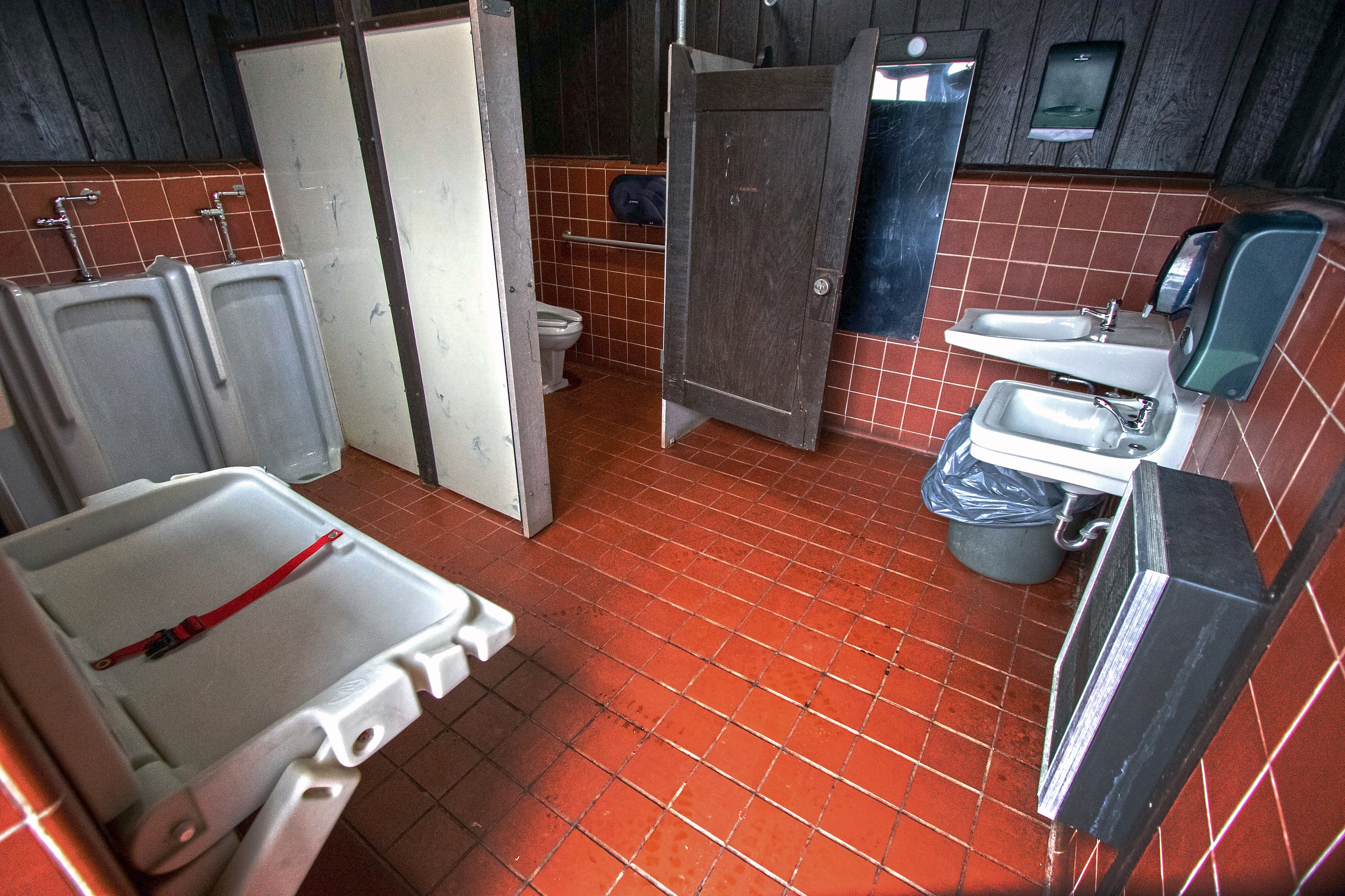 Brown tiled room with changing table and urinals (left), sinks (right), and toilet stall in between.