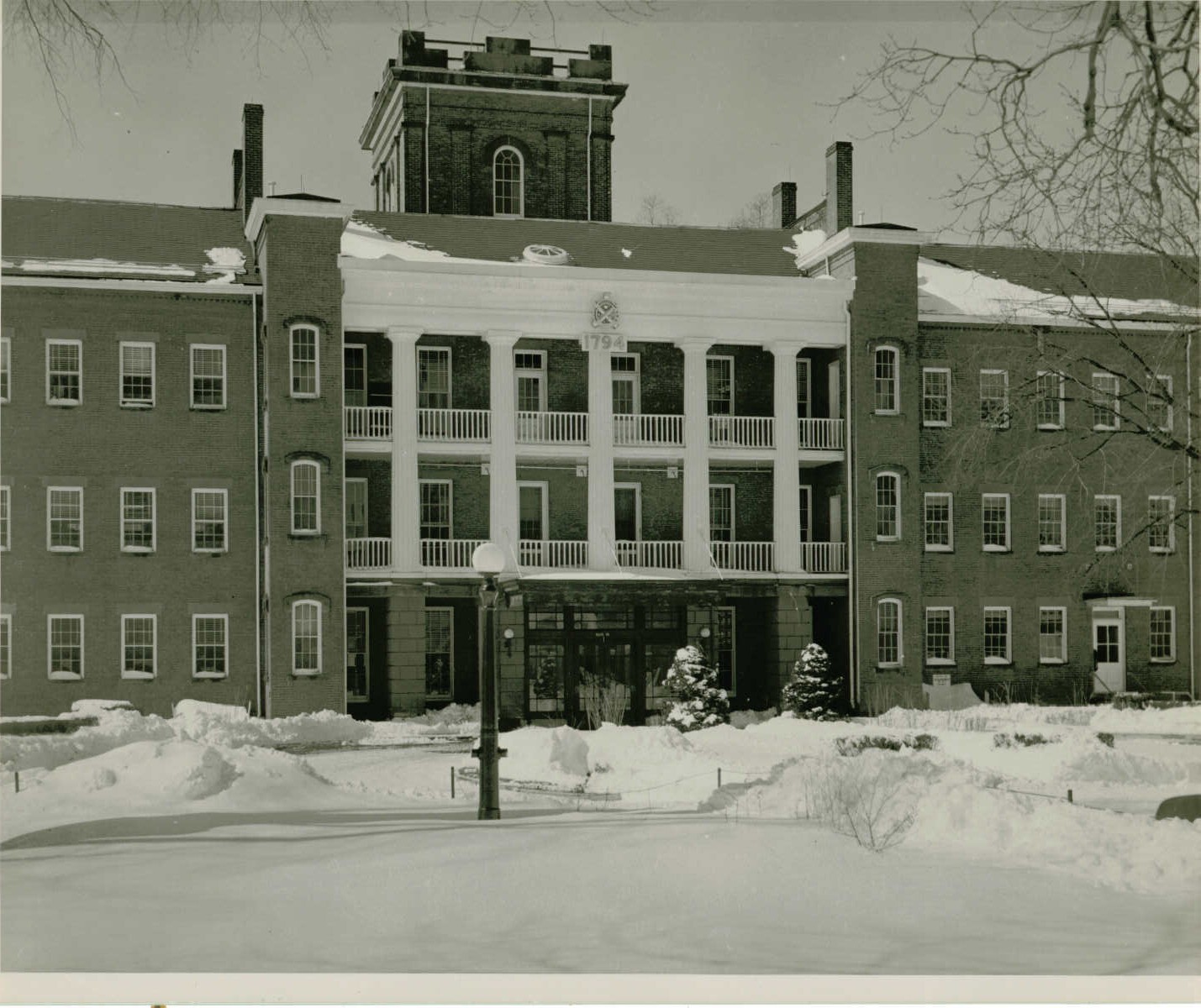 Black and white photo of a large brick building with a second and third story balcony above the entrance. A tower can be seen in the background. An insignia and the year “1794” hangs above the third story balcony. Snow covers the ground. Some snow has been moved off a driveway. A lamppost with a globe light is in the foreground.