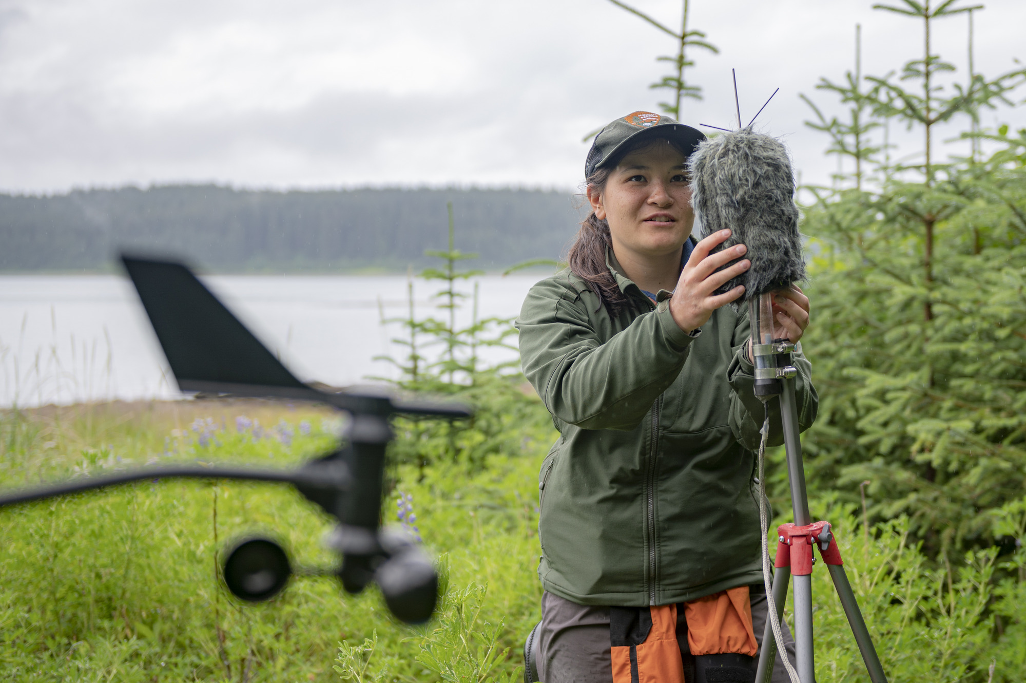 A park ranger scientist handles a microphone windscreen while standing among green vegetation with a waterway in the background.