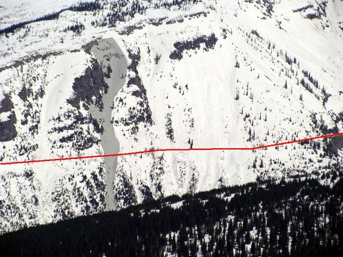 A steep, snowy mountain slope with avalanche paths on its flank is cross-cut by a road.
