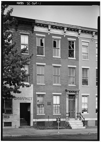 A photo of rowhouses in black and white. The house in the center of the photograph is the focus. It was shattered windows and is clearly rundown.
