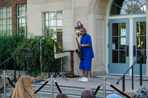 An African American man can be seen in the center of the screen playing a saxophone. A crowd of seated onlookers can be seen in the foreground. An early 20th century school is visible in the background.