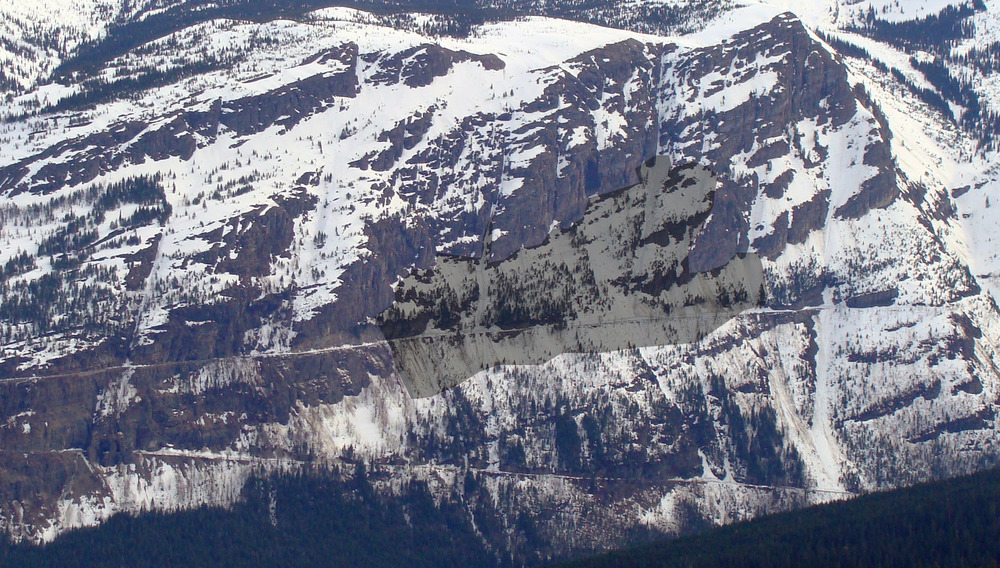 A wide avalanche path runs off of the steep, snowy mountain slopes above a road.