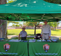 Two people at two green tables with NPS arrowhead and text, set with various items, under a green canopy, buildings, grass and trees in the background.