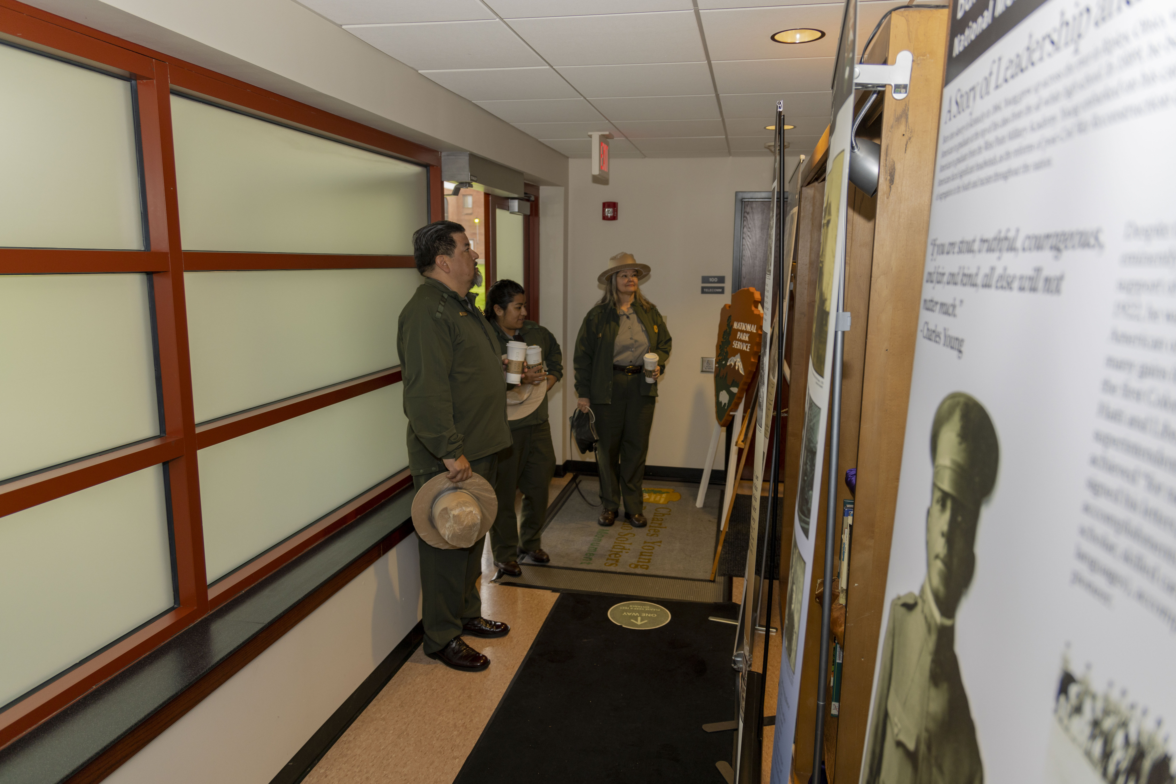 A man in a green uniform holds a tan flat hat and faces a wall of displays to read them.