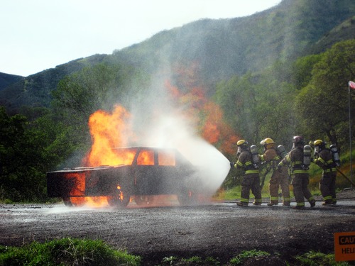 Four firefighters wearing full gear advance pour water from a hose to douse a vehicle on fire 