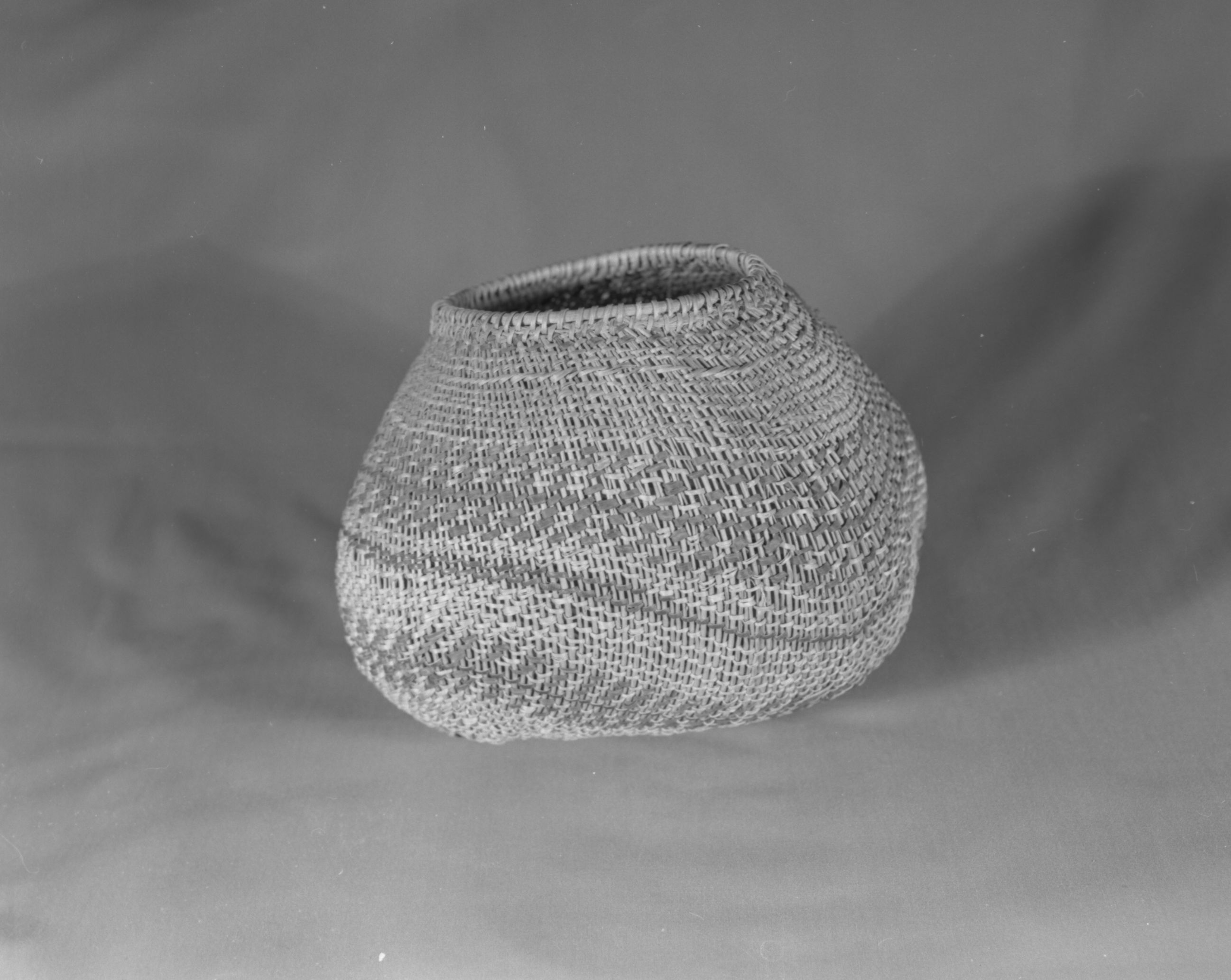 Private Collection. Object: Miwok basket. Dimensions:H-8" x L-9" x W-7 1/2" (maximum) mouth 5". Materials: Scraped shoots, split shoot (probably Ceanothus cuneatus), redbud, bracken fern root, black cloth & red cloth (now white). Technology: Cradle board hood start utilizing cloth wefts. Body of basket in spaced rows of diagonal twining 1/8 to 1/4" apart, one row three strand twining one inch below selvedge, 3 short rows of twining apparently used as a filler on one end of basket. Rim finish of rods folded to left and coiled with small bundle of rods coiled in a 2" section to even out contour. Rim stick attached with aid of an awl. Collection History: From collection of Florence Hill McCullough, daughter of Thomas Hill purportedly from Hill's studio. Photographed by Michael Dixon. Catalogued by Craig Bates and Barbara Beroza. Yosemite Collections, National Park Service.