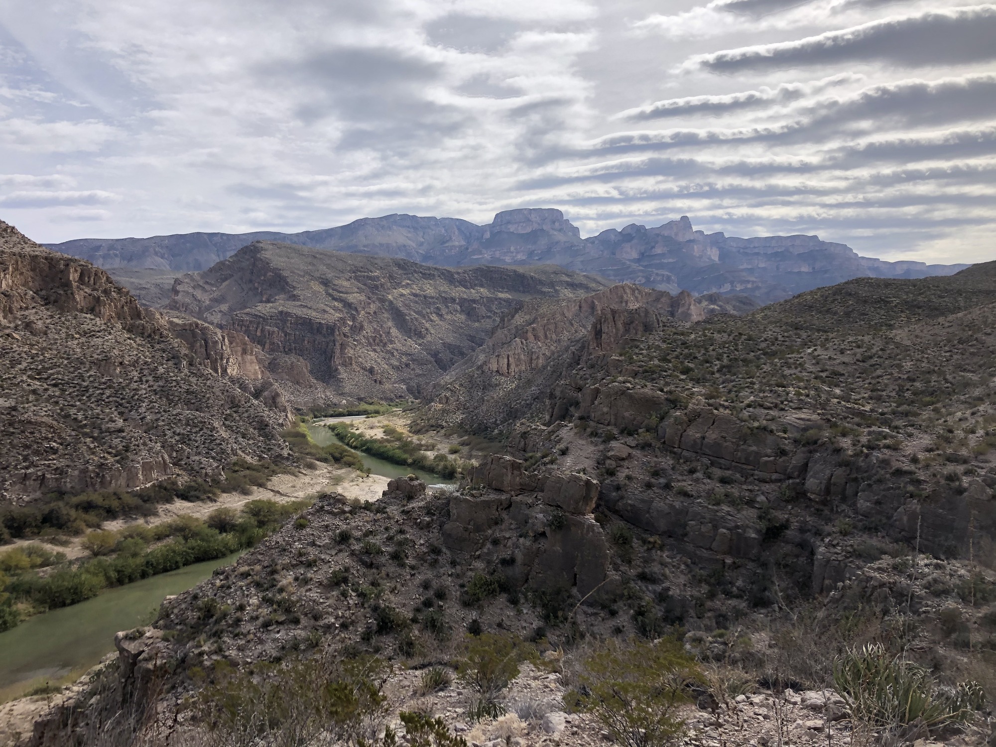 A view of the Rio Grande looking down from Marufo Vega. The river is green and cuts through jagged, beige and brown limestone hills. The Maderas del Carmen is visible in the background, and the sky is filled with gray altostratus clouds.