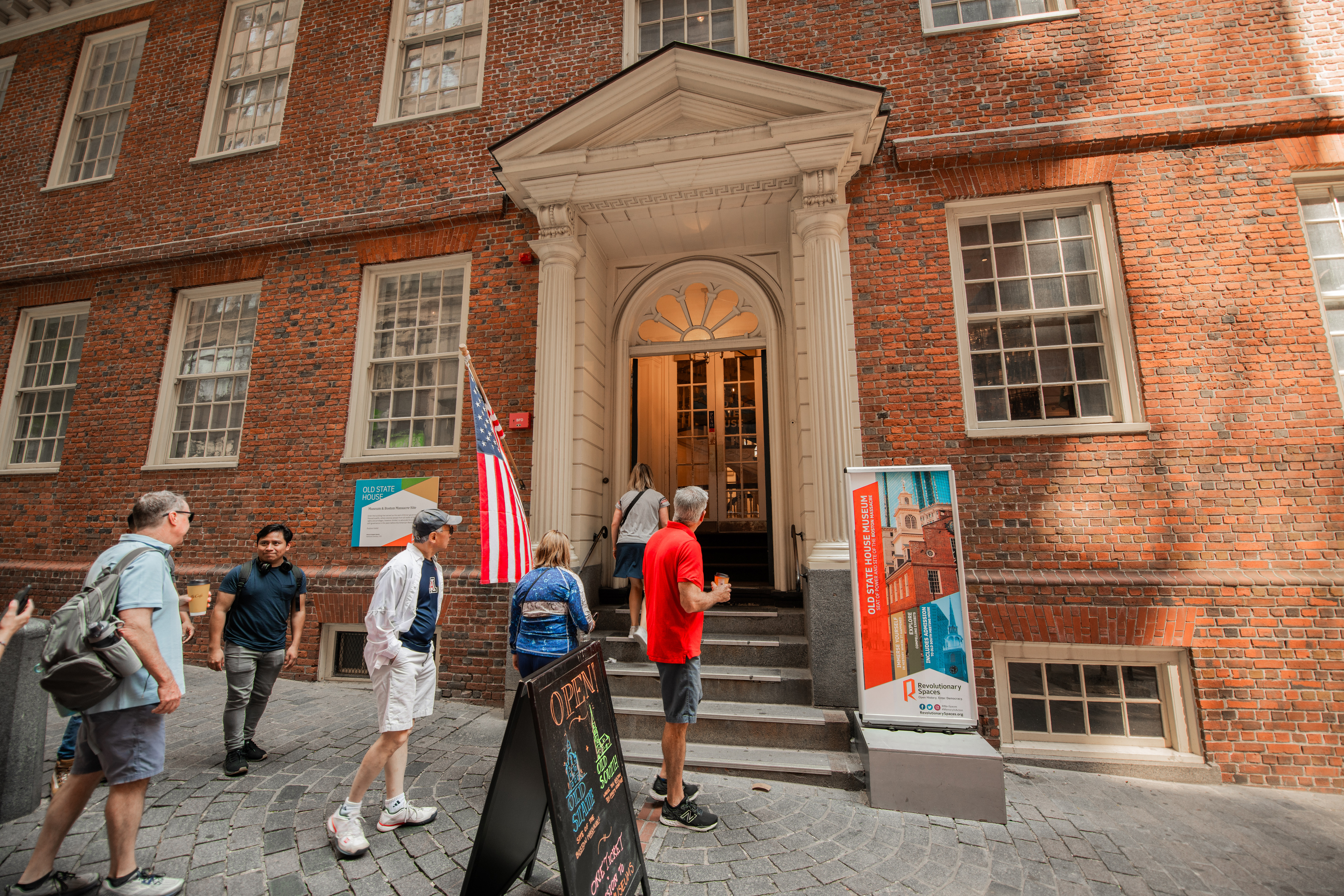 A group of visitors walk up to the entrance of the Old State House, a red brick building. The entrance is marked by two white columns with a pediment on top and has eight stairs which leads visitors through a white arched doorway. 