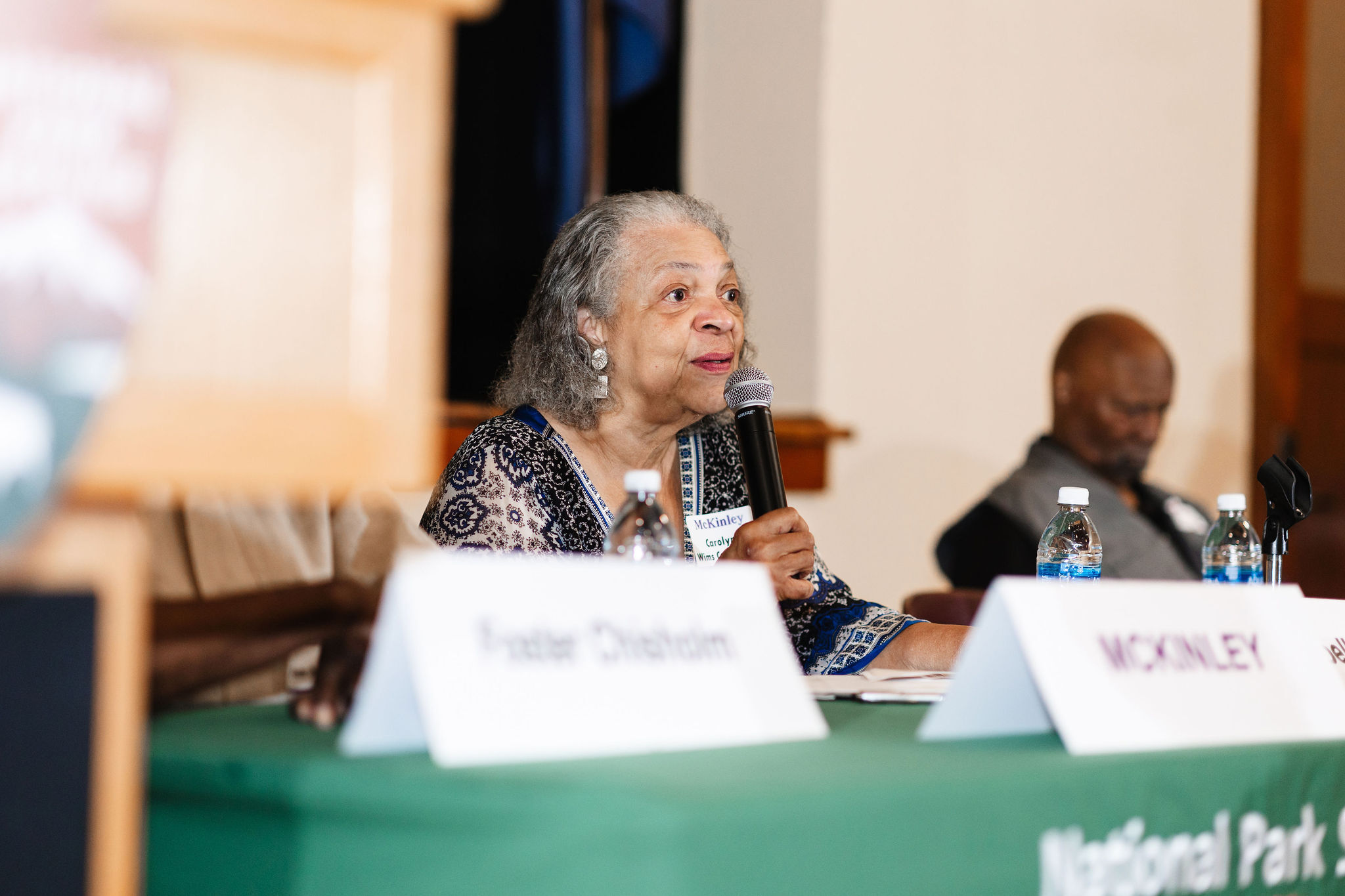 An older african american woman in a blue and black spotted dress speaks into a microphone while seated behind a green table